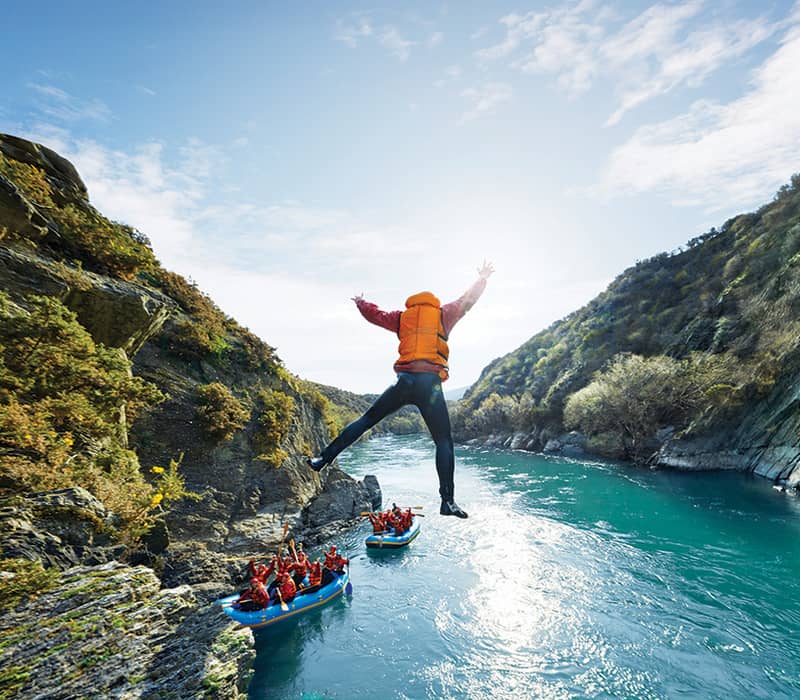 A cliff jumper leaps into the river above two waiting raft crews in a deep canyon gorge. Credit: RealNZ