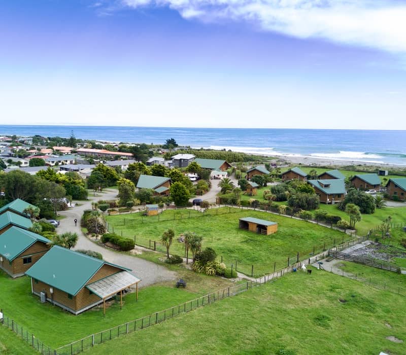 An aerial view of a coastal holiday park featuring several green-roofed timber cabins spaced around grassy paddocks near the ocean. Credit: Shining Star, Hokitika