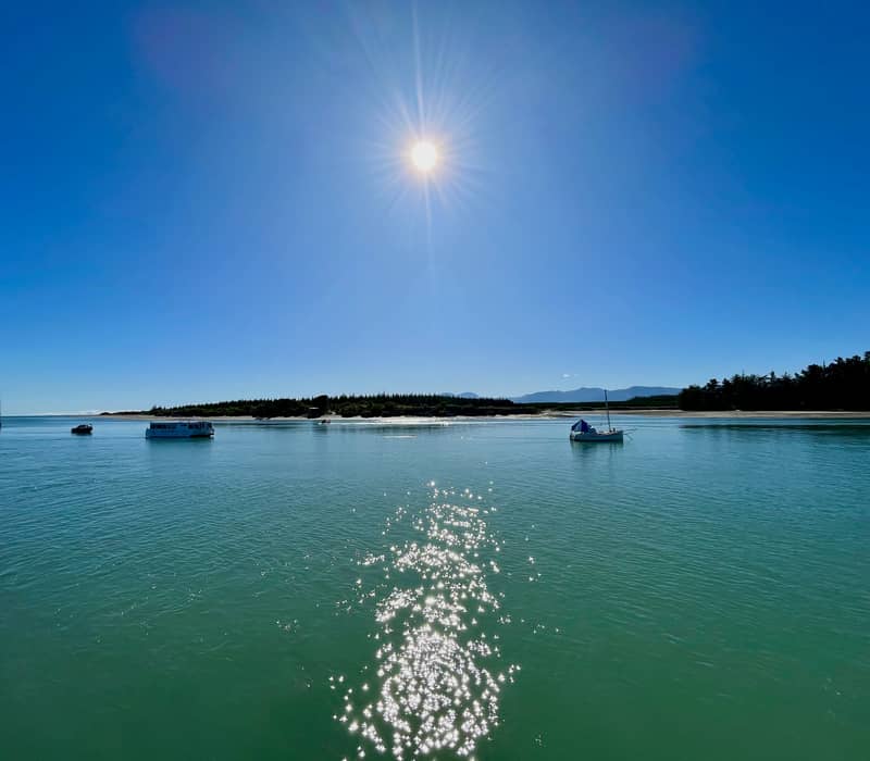 A bright, wide-angle shot from Māpua Wharf looking across the glittering turquoise water of the Waimea Estuary under a blazing high sun, showing anchored sailboats and a water taxi heading toward Rabbit Island. Credit: Richard lumborg - Unsplash
